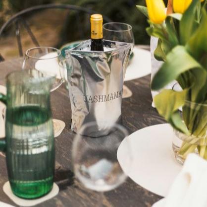 A table set with washable paper placemats is shown. On the table is a glass vase containing yellow tulips, and a silver metallic wine cooler holding a bottle of wine. The UASHMAMA name is printed on the cooler.