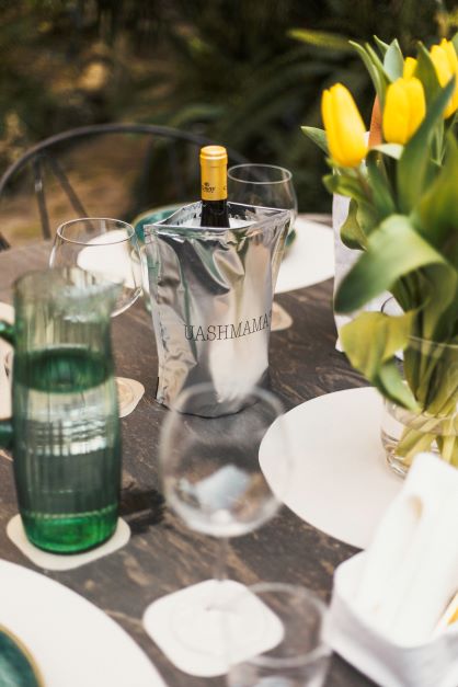 A table set with washable paper placemats is shown. On the table is a glass vase containing yellow tulips, and a silver metallic wine cooler holding a bottle of wine. The UASHMAMA name is printed on the cooler.